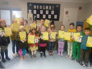 Uczniowie nagrodzeni i wyróżnieni w konkursie pozują do wspólnego zdjęcia trzymając w dłoniach otrzymane dyplomy. Dzieci stoją na szkolnym korytarzu. W tle na ścianie za uczniami wisi gazetka zatytułowana "WIOSNA !".