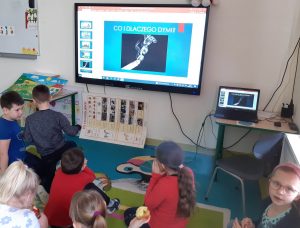 Uczniowie oddziału przedszkolnego siedzą na dywanie i oglądają prezentacje wyświetloną na monitorze zamocowanym na ścianie. Na monitorze wyświetlony jest slajd z napisem "CO I DLACZEGO DYMI?" oraz zdjęciem dymiącego papierosa umieszczonym poniżej.