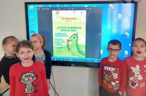 Uczniowie oddziału przedszkolnego stoją przy monitorze umieszczonym na ścianie. Na monitorze wyświetlony jest plakat z napisem " Ta placówka realizuje Program Przedszkolnej Edukacji Antytytoniowej "CZYSTE POWIETRZE WOKÓŁ NAS" Serdecznie zapraszamy przedszkolaki w wieku 6 lat oraz ich rodziców do udziału w programie Dyrekcja i pracownicy przedszkola oraz Państwowy Powiatowy Inspektor Sanitarny w Toruniu." Przy tekście umieszczona jest grafika zielonego smoka wąchającego kwiatki.