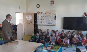 Uczniowie oddziału przedszkolnego siedzą na dywanie w swojej sali. Na przeciwko nich stoi Pan Leśiczy.
