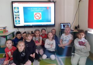 Uczniowie oddziału przedszkolnego siedzą na dywanie. Nad nimi zawieszony jest monitor na którym wyświetlone jest hasło "Co się dzieje, gdy ludzie palą papierosy ?" Pod hasłem jest umieszczony znak zakazu palenia.