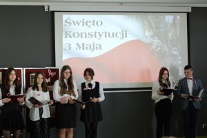 Po lewej stronie stoją w rzędzie cztery uczennice trzymające przed sobą sztywne podkładki. Po prawej stronie stoi dwoje uczniów prowadzących uroczystość. w tle na rzutnkinku wyświetla się zdjęcie polskiej flagi z napisem Święto Konstytucji 3 Maja