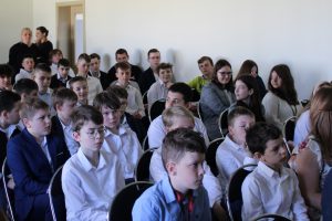 Zdjęcie siedzących na widowni uczniów. Dzieci siedzą na ustawionych w rzędach krzesłach. Ubrani sa w stroje galowe.