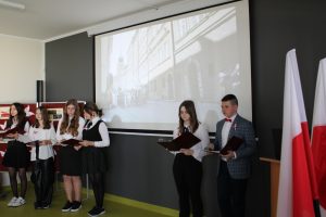 Uczniowie klasy ósmej prowadzących apel stoja na tle rzutnika z wyświetlonym budynkiem. Po prawej stronie stoi dwójka prowadząca spotkanie, a po lewej cztery uczennice ich wspierające.