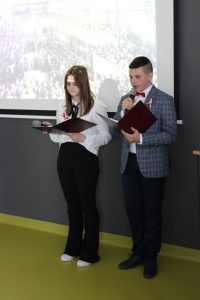 Dwoje uczniów klasy ósmej prowadzących apel syoi obok siebie trzymając mikrofony oraz podkładki z tekstem .
