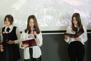 Trzy uczennice klasy ósmej w strojach galowych stoją na tle rzutnika. Dziewczynka stoją po środku trzyma w dłoniach mikrofon.