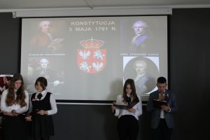 Na tle rzutnika na którym wyświetla się napis "KONSTYTUCJA 3 MAJA 1791 R." oraz zdjęcia mężczyzn którzy przyczynili się do powstania konstytucji stoi czworo uczniów klasy ósmej. Dwoje po prawej stronie i dwie dziewczynki po lewej.