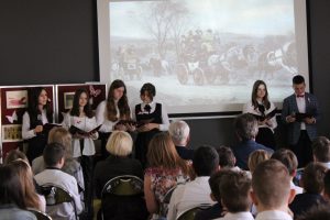 Na tle rzutnika, na którym wyświetla się zdjęcie jadącego powozu i idących ludzi stoją uczniowie klasy ósmej. Po prawej stronie dwójka prowadzących, po lewej cztery uczennice. Przed nimi widać siedzącą widownię.