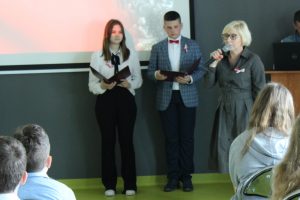 Pani Dyrektor podczas przemówienia na apelu. W dłoni przed sobą trzyma mikrofon. W tle widać dwoje uczniów prowadzących apel a z przodu głowy widowni.