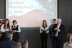 Wójt Gminy Łysomice podczas przemówienia na apelu. W dłoni przed sobą trzyma mikrofon. W tle widać dwoje uczniów prowadzących apel oraz ich dwie koleżanki, a z przodu głowy widowni.