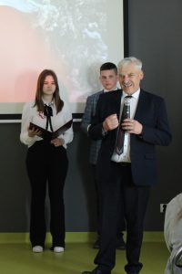 Wójt Gminy Łysomice podczas przemówienia na apelu. W dłoni przed sobą trzyma mikrofon. W tle widać dwoje uczniów prowadzących apel .