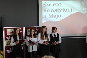 Cztery uczennice klay ósmej stoją w jednym rzędzie. Dziewczynki w strojach galowych, trzymają przed sobą bordowe podkładki. Za nimi na rzutniki wyświetla się flaga polski oraz napis "ŚWIĘTO KONSTYTUCJI 3 MAJA".