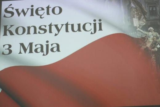 Na powiewającym fragmencie flagi Polski umieszczony napis "Święto Konstytucji 3 Maja". W prawym rogu gdzie flaga zanika widać fragment obrazu na którym jeden mężczyzna jest podnoszony w górę przez innych.