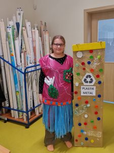 Uczennica klasy piątej pozuje do zdjęcia w wykonanym przez siebie stroju z recyklingu. Obok niej stoi tekturowa tablica w kształcie kosza na śmieci z przyczepionym napisem "PLASTIK METAL" , do której przyklejono plastikowe naklejki i butelki.
