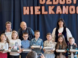 Uczniowie nagrodzeniu w konkursie pozują do wspólnego zdjęcia. Dzieci stoją w jednym rzędzie trzymając w dłoniach prostokątne pudełka- otrzymane nagrody. Za nimi stoi troje dorosłych.