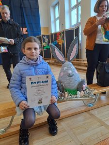Uczennica siedzi na ławeczce w kurtce. Przed sobą trzyma otrzymany dyplom a bok niej ustawiona jest jej praca - zajączek wielkanocny.