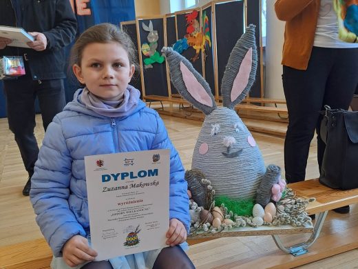 Uczennica siedzi na ławeczce w kurtce. Przed sobą trzyma otrzymany dyplom a bok niej ustawiona jest jej praca - zajączek wielkanocny.