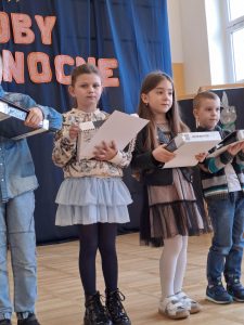 Troje uczniów nagrodzonych w konkursie stoi obok siebie w rzędzie. Dzieci trzymają w dłoniach nagrody - białe pudełka.