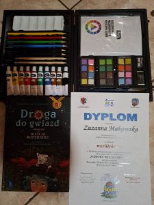 Zdjęcie otrzymanych przez uczennicę nagród oraz dyplomu. Ułożone obok siebie w prostokącie. U góry przybory domalowania a pod spodem dyplom Zuzanny Makowskiej za wyróżnienie w konkursie oraz drugi za udział z napisem "Droga do gwiazd opowieść o Mikołaju Koperniku".