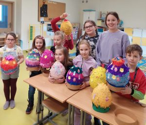 Uczniowie klasy pierwszej wraz z wolontariuszami z klasy piątej pozują do wspólnego zdjęcia stojąc za stołem, na którym ułożone są wspólnie wykonane duże pisanki. Jeden z wolontariuszy podnosi w górę jedną z pisanek zasłaniając sobie nią twarz.