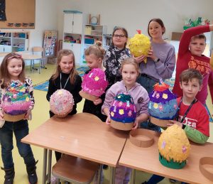 Uczniowie klasy pierwszej wraz z wolontariuszami z klasy piątej pozują do wspólnego zdjęcia stojąc za stołem, na którym ułożone są wspólnie wykonane duże pisanki. Część dzieci również trzyma swoje pisanki w dłoniach.