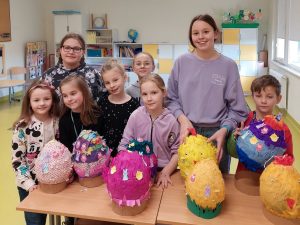 Uczniowie klasy pierwszej wraz z wolontariuszami z klasy piątej pozują do wspólnego zdjęcia stojąc za stołem, na którym ułożone są wspólnie wykonane duże pisanki.