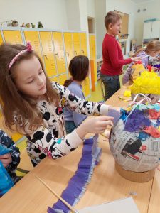 Uczennica klasy pierwszej podczas ozdabiania swojej pisanki. Dziewczynka stoi przy stole i trzyma w dłoniach długi pasek niebieskiej krepy, którym ozdabia jajko. W tle widać dwójkę pracujących wolontariuszy.