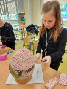 Uczennica klasy pierwszej stoi przy stoliku, na którym ustawione jest duże jajko wykonane z papieru. Dziewczynka je obkleją papierowymi serwetkami w różowe kropki.