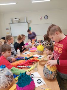 Uczniowie klasy pierwszej i wolontariusze przy wspólnej pracy. Dzieci siedzą lub stoją przy długim stole, na którym ustawione są duże jajka wykonane z papieru oraz przybory plastyczne. Dzieci ozdabiają swoje pisanki.