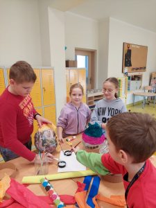 Wolontariusze i uczniowie klasy pierwszej podczas pracy przy wielkanocnych pisankach. Dzieci stoją przy stole, na którym ustawione są papierowe jajka oraz przybory plastyczne.