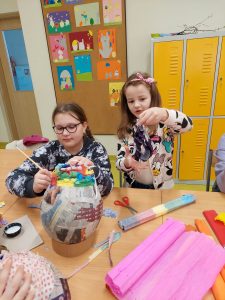 Uczennica klasy pierwszej oraz wolontariusza z klasy piątej wspólnie pracują nad jedną z pisanek. Wolontariuszka siedzi przy stole trzymając w dłoni pędzelek oraz malowaną pisankę. Obok stoi uczennica trzymając w dłoniach kawałki papieru. Na stole obok jajka leżą przybory plastyczne i kolorowe krepy.