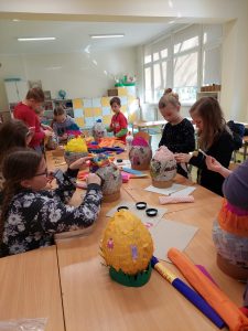 Uczniowie klasy pierwszej i wolontariusze przy wspólnej pracy. Dzieci siedzą lub stoją przy długim stole, na którym ustawione są duże jajka wykonane z papieru oraz przybory plastyczne. Dzieci ozdabiają swoje pisanki.