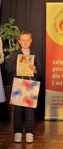Nagrodzony uczeń pozuje do zdjęcia z otrzymanym dyplomem i nagrodą. Chłopiec uśmiecha się.