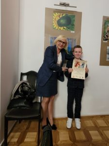Pani dyrektor pozuje wraz z nagrodzonym uczniem do zdjęcia. chłopiec trzyma przed sobą dyplom. Na ścianie za nimi wiszą nagrodzone prace.