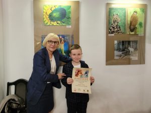 Pani dyrektor pozuje wraz z nagrodzonym uczniem do zdjęcia. chłopiec trzyma przed sobą dyplom. Na ścianie za nimi wiszą nagrodzone prace.