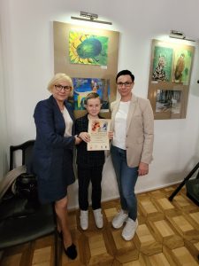 Pani Dyrektor pozuje wraz z nagrodzonym uczniem oraz jego mamą do zdjęcia. Chłopiec trzyma przed sobą dyplom. Na ścianie za nimi wiszą nagrodzone prace.