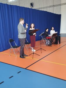 Dwójka muzyków stoi trzymając mikrofony. w tym samym rzędzie siedzą jeszcze dwie Panie za kontuarem z kibordem.