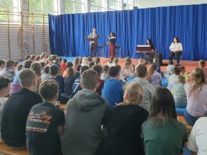 Na przeciwko uczniów siedzących na długich ławkach i materacach stoi dwoje muzyków. Obok nich siedzą kolejne dwie Panie a przed jedną z nich stoi na stelażu kaybord.