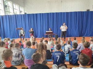 Na przeciwko uczniów siedzących na długich ławkach i materacach siedzi troje muzyków. Obok nich stoi Pani z mikrofonem.