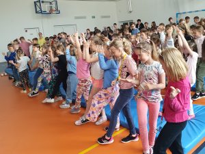 Uczniowie szkoły podczas audycji muzycznej. Dzieci w tańcu podnoszę w górę ręce i nogi.