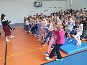Uczniowie szkoły podczas audycji muzycznej. Dzieci w tańcu podnoszę w górę ręce i nogi. Na przeciwko nich stoją dwie Panie również z podniesionymi w górę nogami.