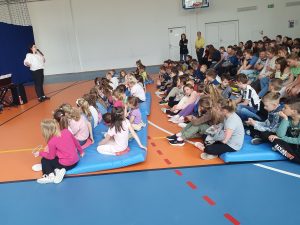 Uczniowie podczas audycji muzycznej siedzą na materacach na sali gimnastycznej. Przed nimi stoi Pani trzymająca przy ustach mikrofon.
