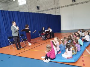 Uczniowie podczas audycji muzycznej siedzą na materacach na sali gimnastycznej. Przed nimi stoi dwójka muzyków a dalej siedzą jeszcze dwie Panie za kontuarem z kibordem.
