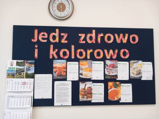 Zdjęcie granatowej tablicy zawieszonej na ścianie z gazetką zatytułowaną "Jedz zdrowo i kolorowo". Pod tytułem gazetki zamieszczono zdjęcia różnych kolorowych potraw wraz z informacji o nich. Z boku po lewej stronie tablicy wisi kalendarz a nad tablicą zegar.