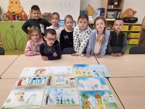 Uczniowie oddziału przedszkolnego pozują do zdjęcia przy dużym stole, na którym ułożone są ich rysunki.