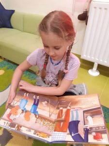 Uczennica oddziału przedszkolnego podczas czytania książki. Dziewczynka przygląda się obrazkom w niej zamieszczonych.