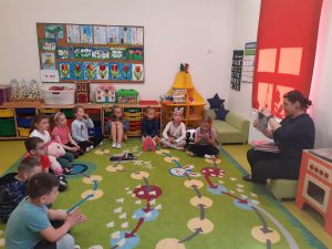 Uczniowie oddziału przedszkolnego siedzą w półkolu na dywanie. Na przeciwko nich siedzi Pani bibliotekarka trzymająca w dłoniach książkę.