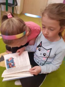 Dwie uczennice oddziału przedszkolnego siedzą na podłodze. Jedna z nich trzyma otworzoną książkę, nad którą pochyla się jej koleżanka.