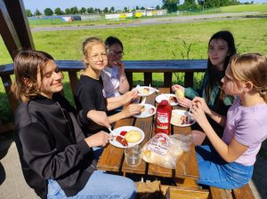 Pięć uczennic siedzi przy stoliku. Przed nimi stoją talerzyki z kanapkami, chleb i ketchup. Dziewczynki spoglądają w stronę zdjęcia.