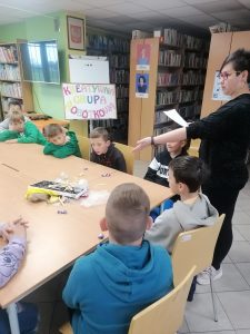 Uczniowie klasy trzeciej siedzą przy dużym stole w gminnej bibliotece. Przy stole stoi Pani bibliotekarka trzymająca w dłoni kartkę i wskazująca drugą ręką leżące na stole plastikowe przedmioty.